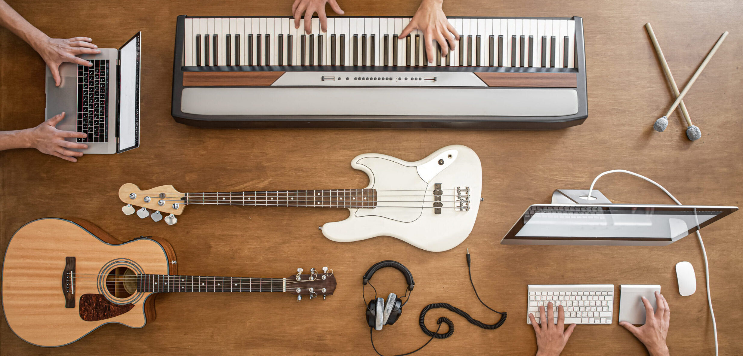 On a wooden table there are musical keys, acoustic guitar, bass guitar, sound mixer, headphones, computer and drum sticks.